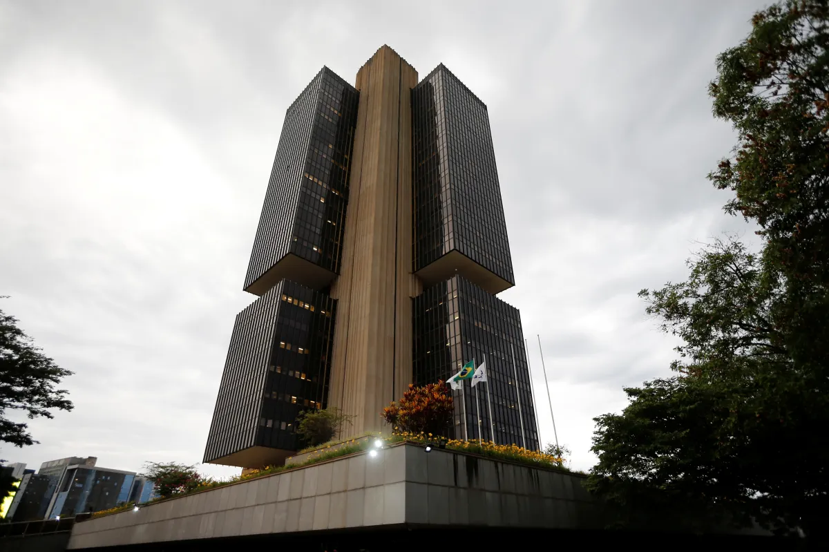 Gráfico ilustrativo com curvas de projeção de inflação para os anos de 2026, 2027 e 2028, ao fundo sede do Banco Central do Brasil em Brasília