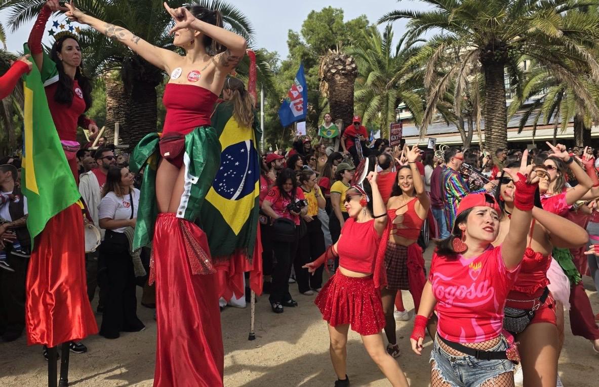 Manifestantes vestidos de vermelho caminham em Barcelona durante ato de apoio à reeleição de Lula, com música e apresentações culturais.